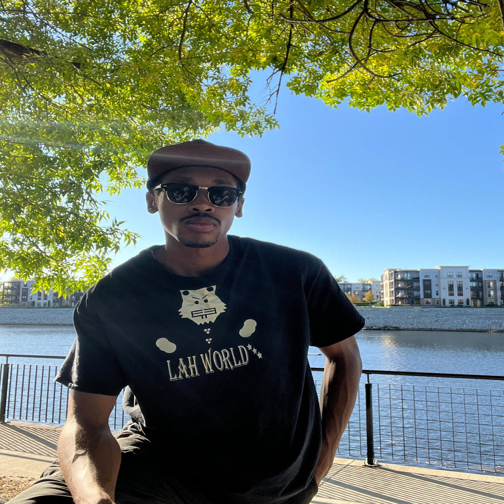 Person wearing a black t-shirt with text and graphics, standing outdoors near trees and water.